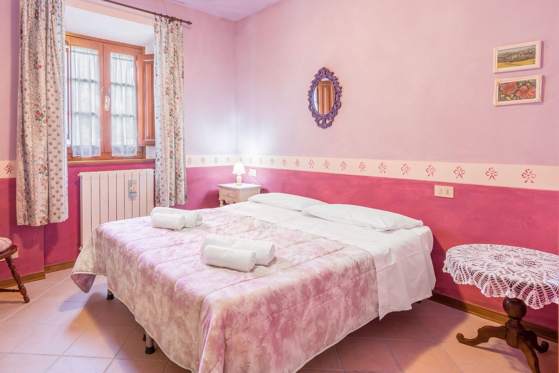 A bedroom with a bed, a nightstand, a window with curtains, a mirror, and a small table with a lace tablecloth.