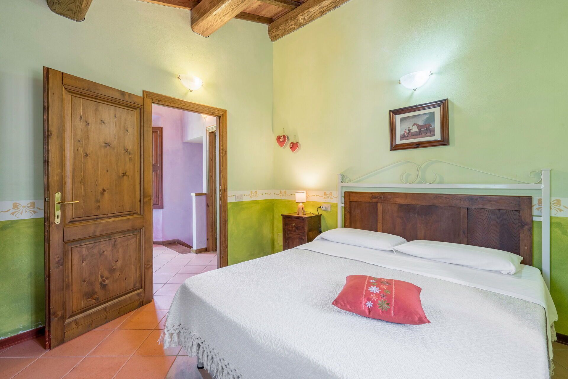 A bedroom with a wooden door, a bed with white bedding and a red pillow, a nightstand with a lamp, and a framed picture on the wall.