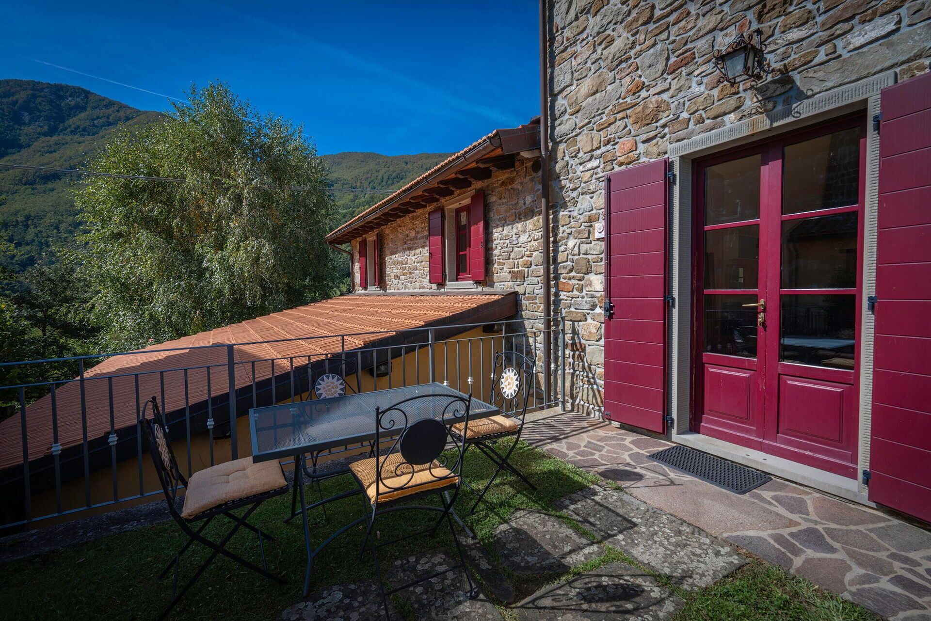 A stone building with red shutters, a balcony with a metal railing, and a table with chairs outside.