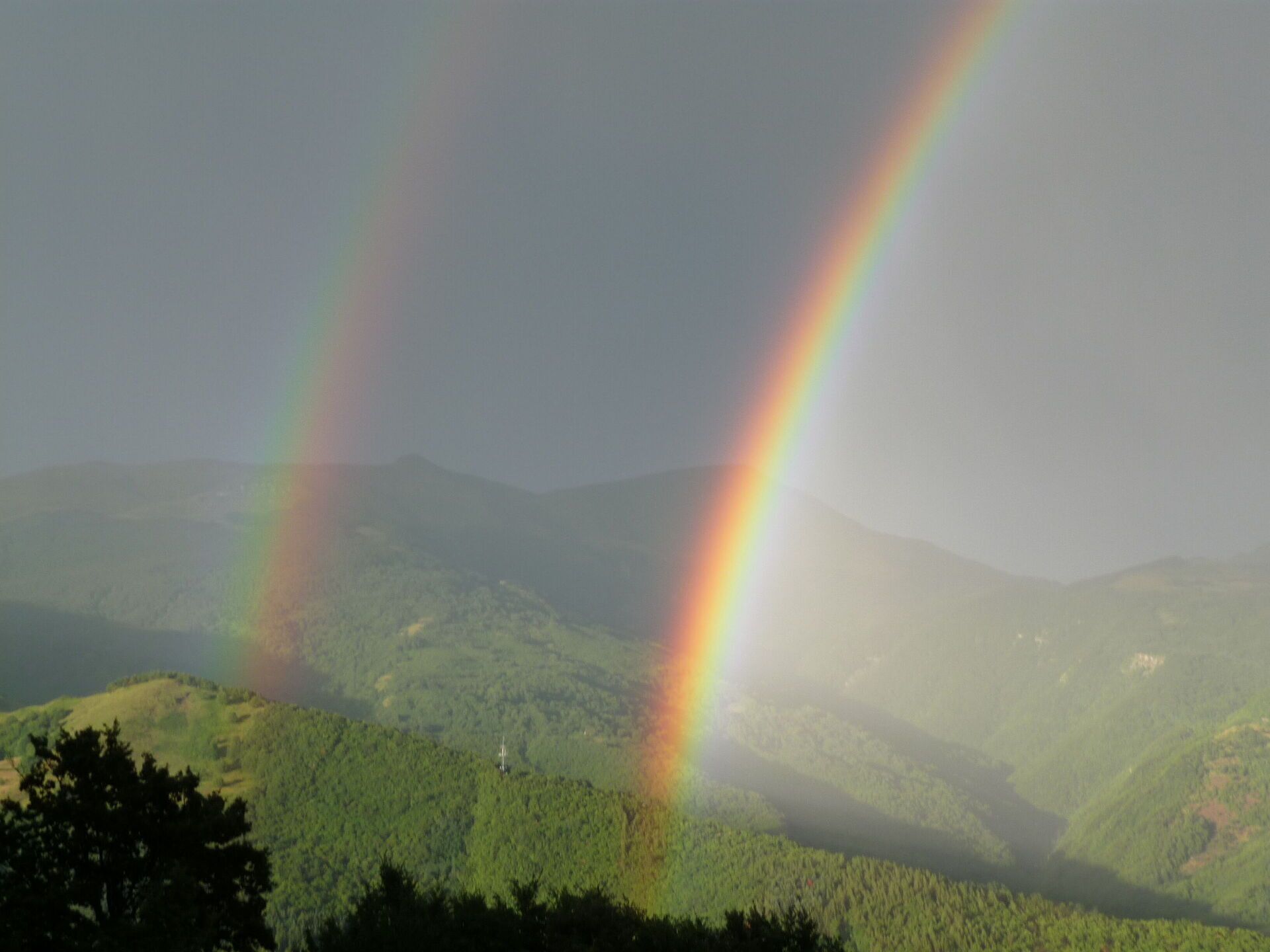 A landscape with two rainbows over a forested mountain range.
