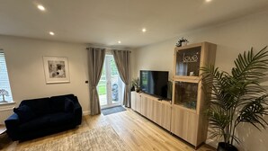 Een woonkamer met een donkerblauwe bank, een flatscreen televisie op een houten kast, een grote potplant en een glazen kastje met de tekst 'love'.