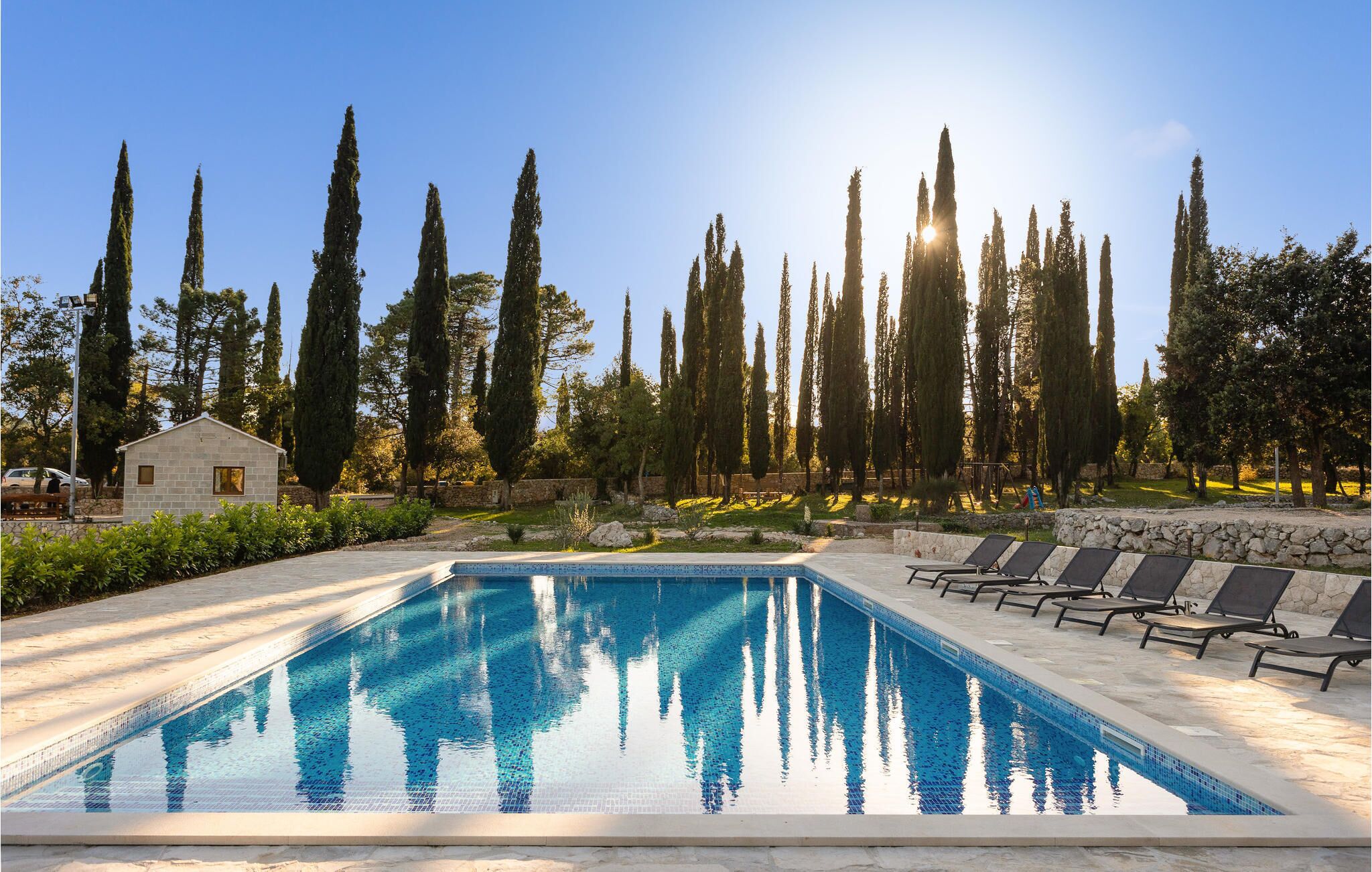 A rectangular swimming pool with clear blue water, surrounded by lounge chairs and a stone wall. Tall cypress trees are visible in the background.