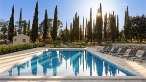 A rectangular swimming pool with clear blue water, surrounded by lounge chairs and a stone wall. Tall cypress trees are visible in the background.
