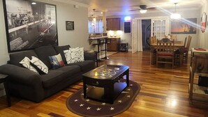 A spacious living room with a wooden floor, a black sofa, a coffee table, and a dining area with a wooden table and chairs. - Wildcatter resting grounds, spacious living (Odessa)