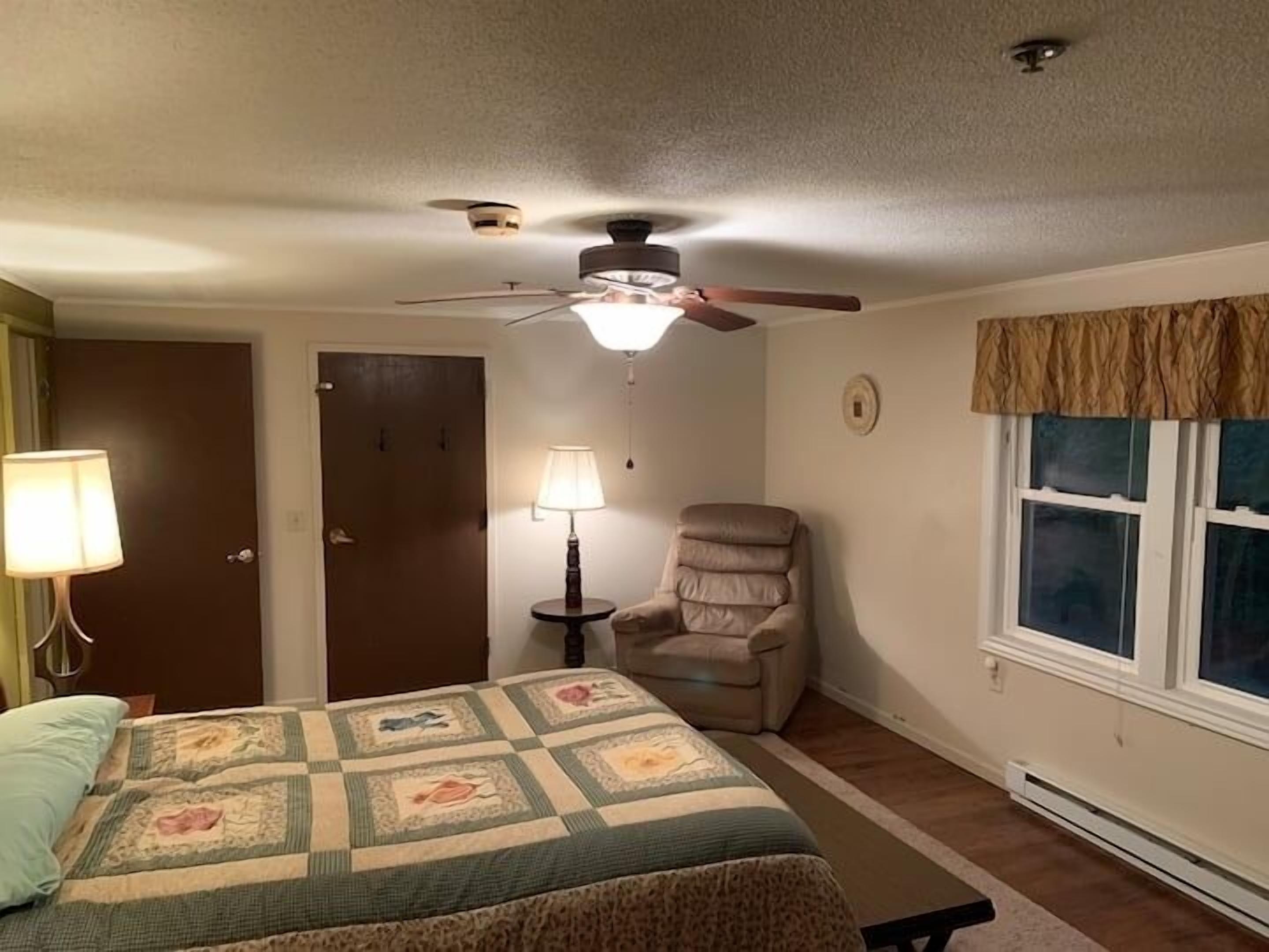 A bedroom with a bed, a chair, a ceiling fan, a window, and a door.