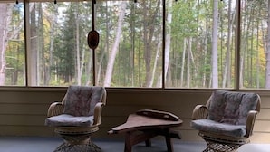 Habitación con dos sillas de mimbre y una pequeña mesa de madera, ventanas amplias con vista al bosque.