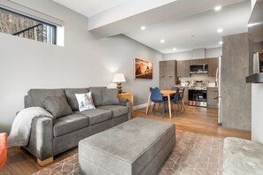 Una sala de estar moderna con un sofá gris, un comedor con sillas azules y una cocina con electrodomésticos de acero inoxidable.