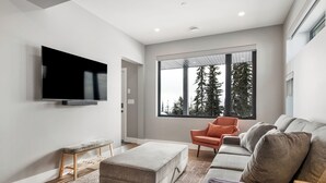 Un salon moderne doté d’un téléviseur à écran plat, d’un canapé gris, d’un fauteuil orange et d’une grande fenêtre donnant sur un paysage arboré.