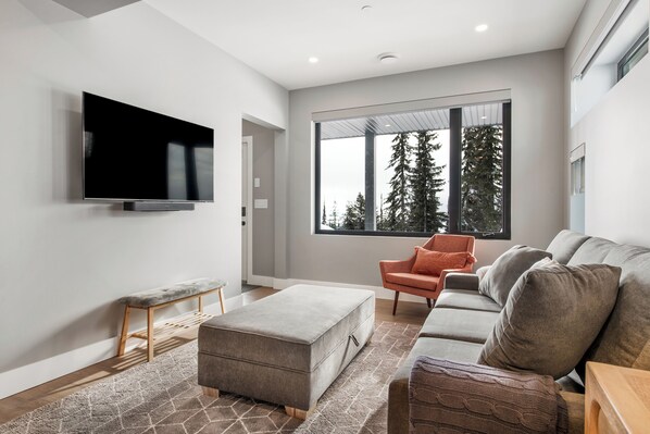 A modern living room with a flat-screen TV, a grey sofa, an orange armchair, and a large window with a view of trees. - Private fully-equipped suite with garage, laundry, and gear room (Silver Star Mountain)