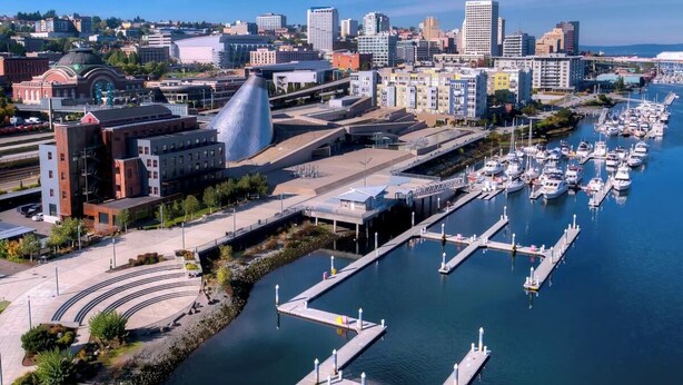 Un port de plaisance avec des bateaux amarrés, un bâtiment moderne doté d’une structure en forme de dôme originale, et un paysage urbain avec divers bâtiments en arrière-plan.