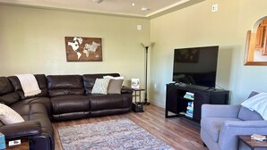 Un salon avec un canapé en cuir marron, un fauteuil gris, un tapis à motifs, un téléviseur à écran plat et une bibliothèque.