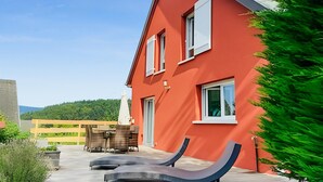 Un edificio rojo con dos sillones, una terraza de madera y vistas a una colina boscosa.
