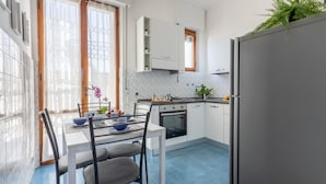 Una cocina con armarios blancos, una mesa de comedor con sillas, un refrigerador y una ventana con cortinas.