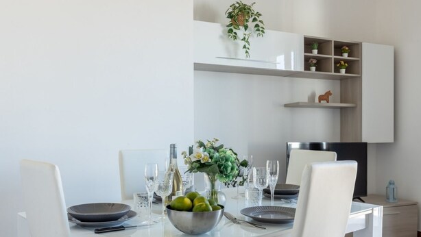Un comedor moderno con una mesa blanca dispuesta para cuatro personas, un frutero y un jarrón con flores.