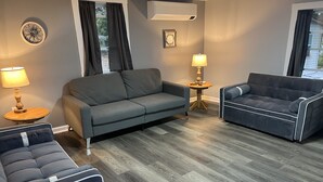 A modern living room with a grey sofa, a blue sofa, and a dark blue armchair. There is a small round table with a lamp, a wall-mounted air conditioner, and a decorative wall piece.