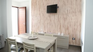 A modern dining area with a wooden wall, a flat-screen TV mounted above the table, and a sliding door leading to another room. - Al Civico 5 - Bright Apartment W/ Private Parking! (Decimomannu)