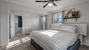 A bedroom with a large bed, a ceiling fan, a window with curtains, a bedside table, and a wall-mounted shelf with a decorative item.