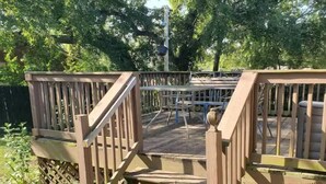 A wooden deck with a staircase leading to a higher level, surrounded by a fence and trees.