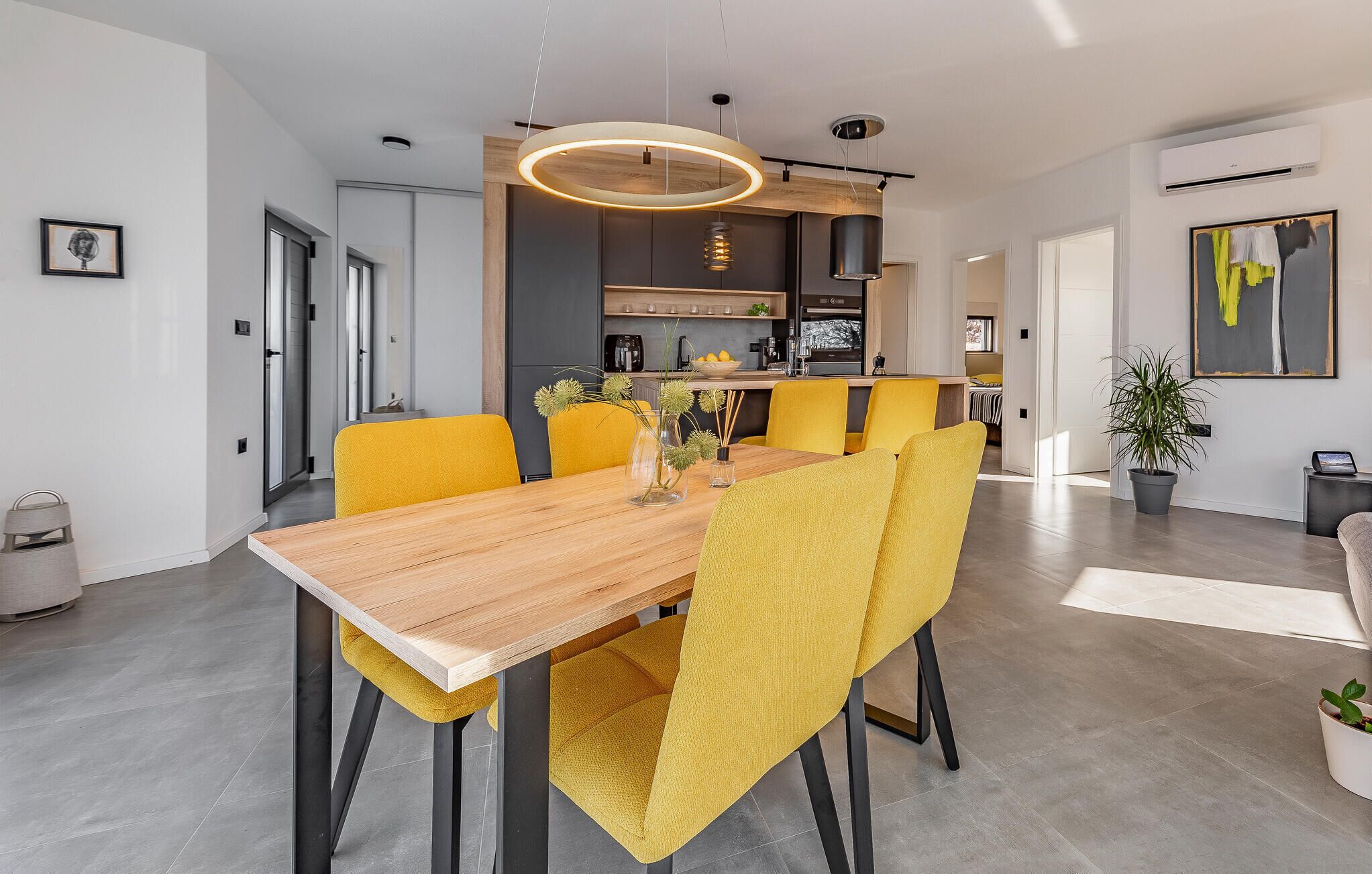 A modern dining area with a wooden table and yellow chairs, a kitchen in the background, and a potted plant on the right.