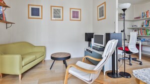 A modern living room with a green sofa, a white chair, a small round table, a desk with a computer, a bookshelf, and framed artwork on the wall. - Appartement Charenton - Welkeys (Paris)