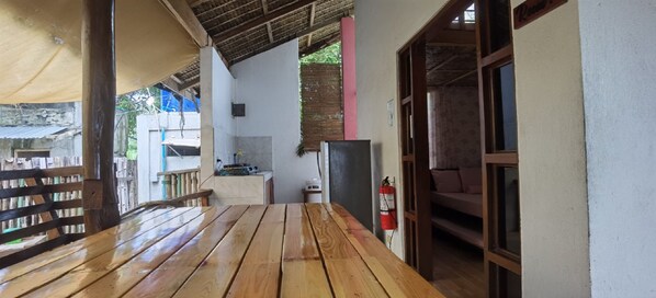 A wooden table and benches in a covered outdoor area with a kitchen in the background.