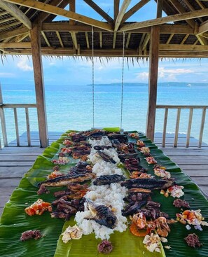 A long table with a variety of dishes, including fish and rice, set up on a wooden deck overlooking the ocean.