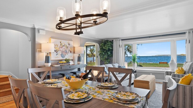 A dining area with a wooden table set for a meal, a chandelier, and a view of the sea through large windows. - Sunshine Cove - Door County (Sturgeon Bay)