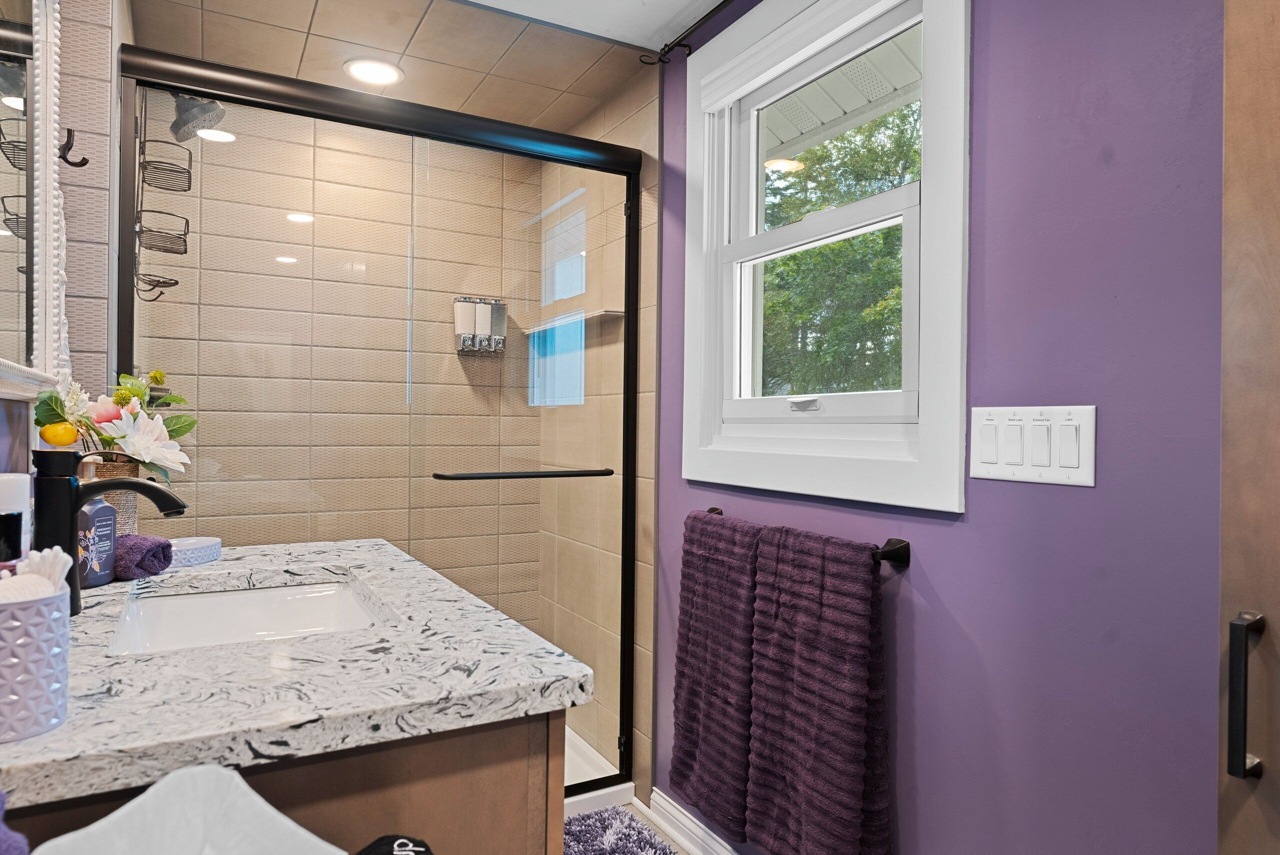 Un baño con ducha, un lavamanos con encimera de mármol, una toalla morada y una ventana con vistas a árboles.