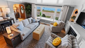 A living room with a sofa, ottoman, armchair, and a fireplace. There is a view of the outdoors through large windows. - Sunshine Cove - Door County (Sturgeon Bay)