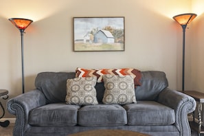 Um sofá de couro cinzento com almofadas estampadas, um quadro emoldurado de um celeiro e duas luminárias de chão.