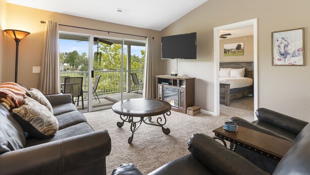 A spacious living room with a sofa, a coffee table, and a flat-screen TV mounted on the wall. There is a sliding glass door leading to a balcony with outdoor furniture.