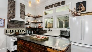 A kitchen with a central island, white cabinetry, a stove, a sink, and a refrigerator. There are wooden shelves above the sink and a sign that reads "COTTON". - The Dollhouse | 3/2 | Walk to Downtown (Natchez)