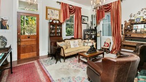 A cozy living room with a sofa, a coffee table, a leather armchair, a wooden cabinet, and a piano. - The Dollhouse | 3/2 | Walk to Downtown (Natchez)
