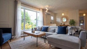 Un salón moderno con un sofá, una mesa de centro y un sillón azul. Hay un ventanal con cortinas, un espejo redondo en la pared y una planta en la mesita de noche.