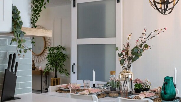 Un comedor con una mesa blanca dispuesta para dos personas, con un jarrón de flores y una jarra verde.