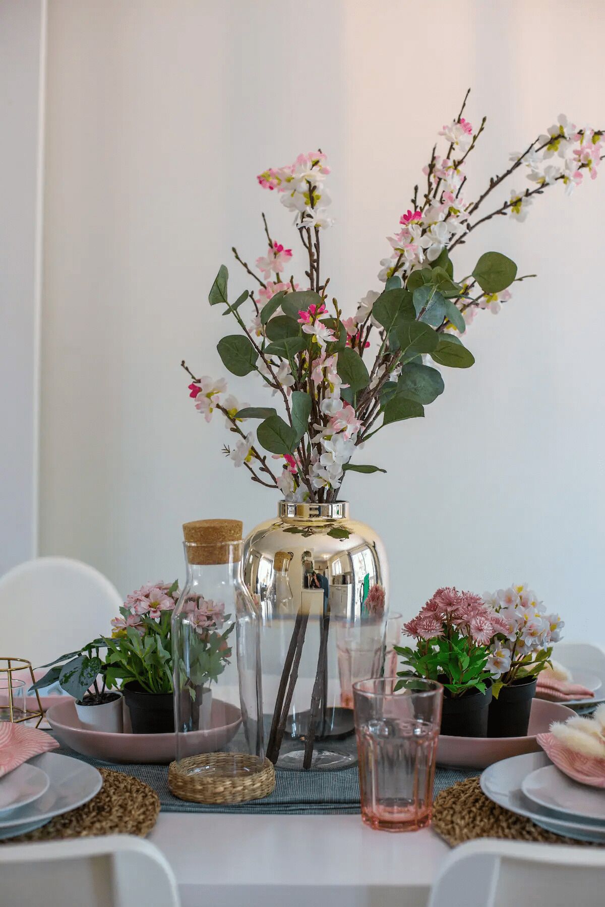 Een gedekte tafel met glazen flessen, een metalen vaas met roze bloemen en een glas water.