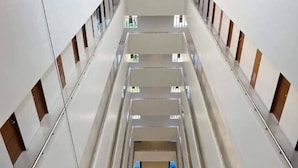 Espacio interior de varios niveles con una escalera central, barandas de vidrio y vistas a un nivel inferior con gente y mobiliario.