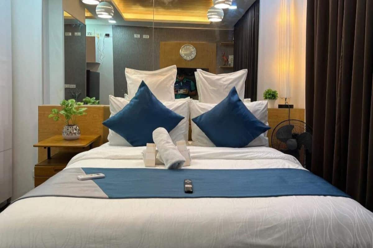 Une chambre d’hôtel moderne avec un grand lit, deux oreillers bleus, une table de chevet avec une plante et un petit ventilateur.