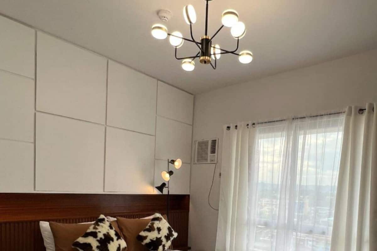 Une chambre à coucher avec un lit, une tête de lit en bois, un luminaire au plafond et une fenêtre avec des rideaux.