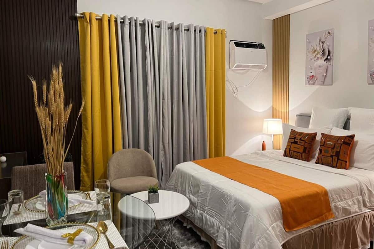 Une chambre d’hôtel avec un lit, une chaise, une petite table, un vase contenant des plantes séchées et un climatiseur fixé au mur.