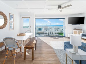 Een ruime woonkamer met een groot raam met uitzicht op een strand, een eethoek met een witte tafel en rieten stoelen, en een moderne tv aan de muur.