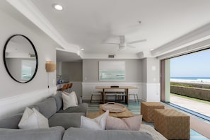 Un salon moderne avec un canapé gris, une table basse en bois et une vue sur la plage, visible à travers une grande fenêtre.