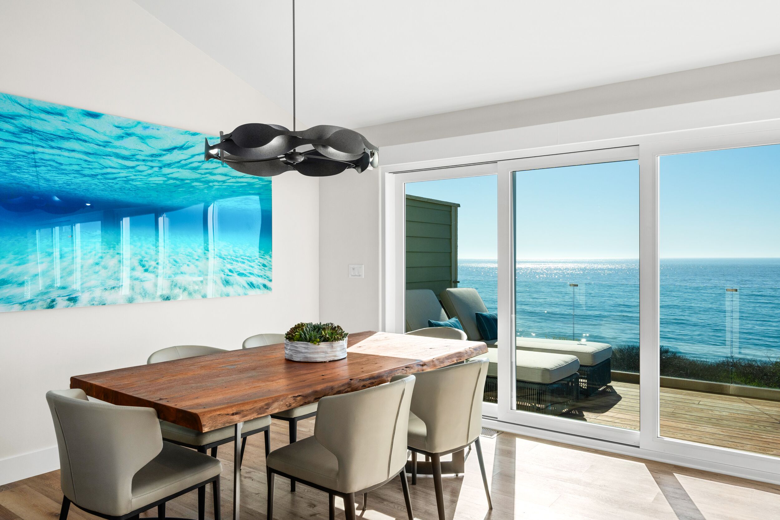 A dining area with a wooden table, four chairs, a potted plant, and a large ocean view through floor-to-ceiling windows.