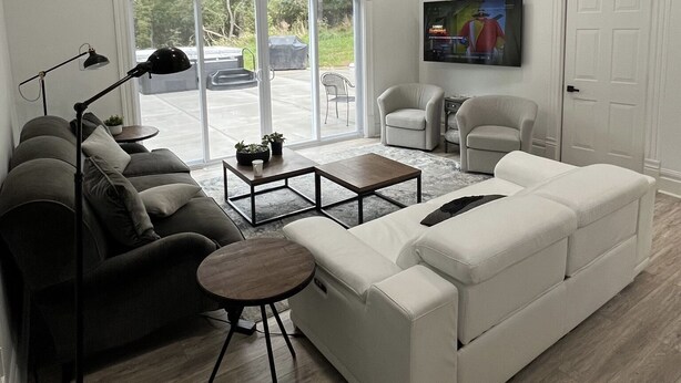A modern living room with a sectional sofa, a small coffee table, and a floor lamp. There is a television mounted on the wall and a sliding glass door leading to an outdoor area with a car and a covered object. - Private Vacation Home with large Hot Tub and Fire Pit -- Sleeps 18 (Gibsonia)