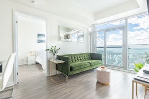 A modern living room with a green sofa, a small side table, a white bedside table, and a large window offering a city view. - Beautiful Luxurious Modern With Breathtaking Views  (Toronto)