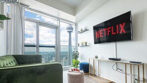A modern living room with a large window, a green sofa, a pink ottoman, a blue chair, a TV mounted on the wall, and a city view.