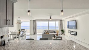 Une cuisine moderne dotée d’un grand îlot central, d’un coin repas avec des chaises, d’un canapé et d’un téléviseur fixé au mur.