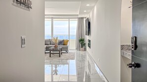 Un intérieur moderne agrémenté d’une pancarte « Bienvenue », un espace de vie avec un canapé et une table basse, une grande fenêtre offrant une vue dégagée et une porte élégante dotée d’une poignée.