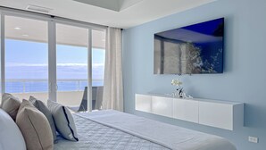 Une chambre avec un grand lit, un téléviseur fixé au mur, une vue sur la mer par la fenêtre et une console blanche.