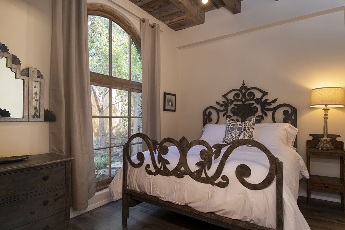 Un dormitorio con un ventanal grande, una cama con cabecero decorado, una mesita de noche con lámpara y un aparador.